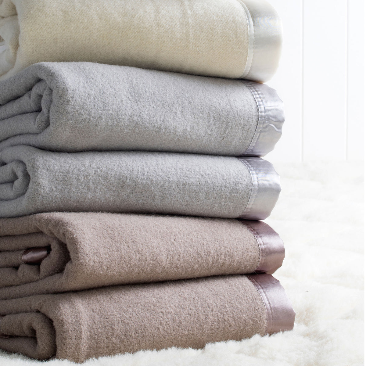 A stack of folded Australian washable wool blankets in ivory, platinum grey, and angora brown, with satin borders.