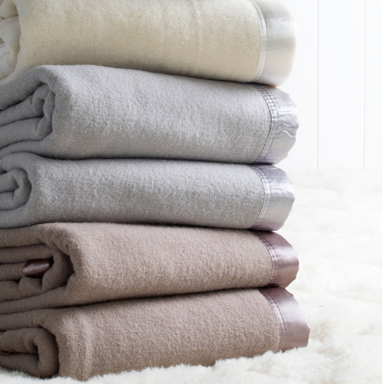 A stack of folded Australian washable wool blankets in ivory, platinum grey, and angora brown, with satin borders.