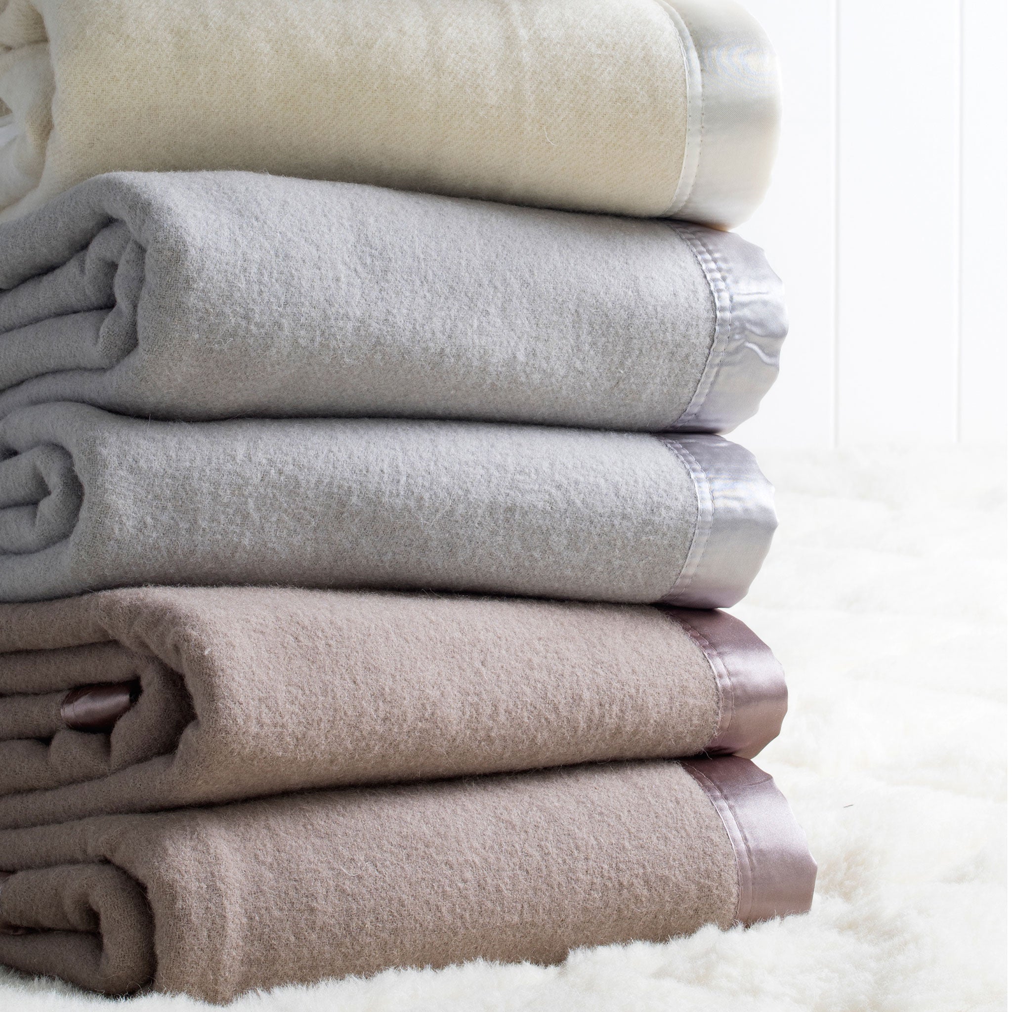A stack of folded Australian washable wool blankets in ivory, platinum grey, and angora brown, with satin borders.