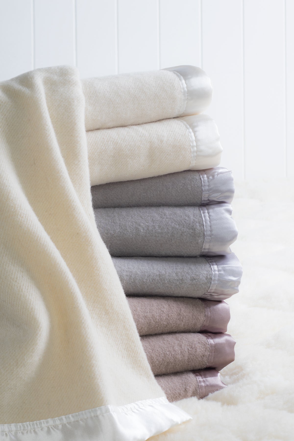 Stack of blankets with satin-bound edges in ivory, platinum grey and angora brown colourways on a woollen underlay, with an ivory blanket draped in the foreground.