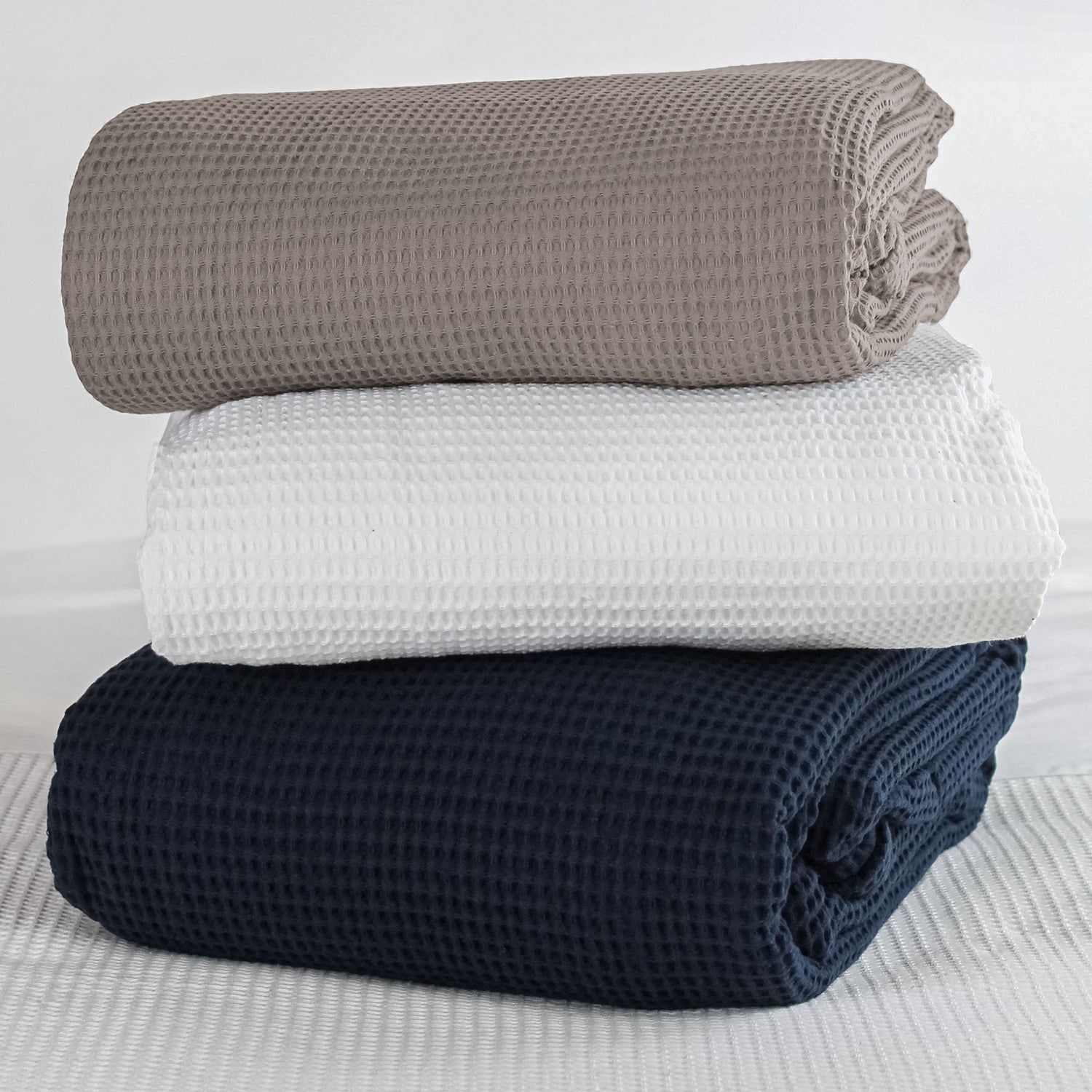 Three folded cotton waffle-weave blankets in latte, white and navy colourways on a bed.