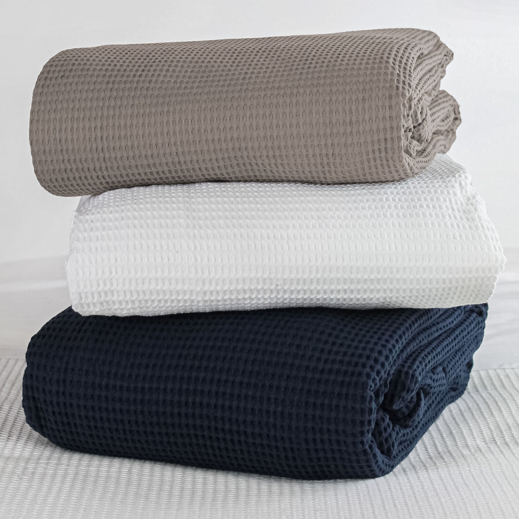 Three folded cotton waffle-weave blankets in latte, white and navy colourways on a bed.