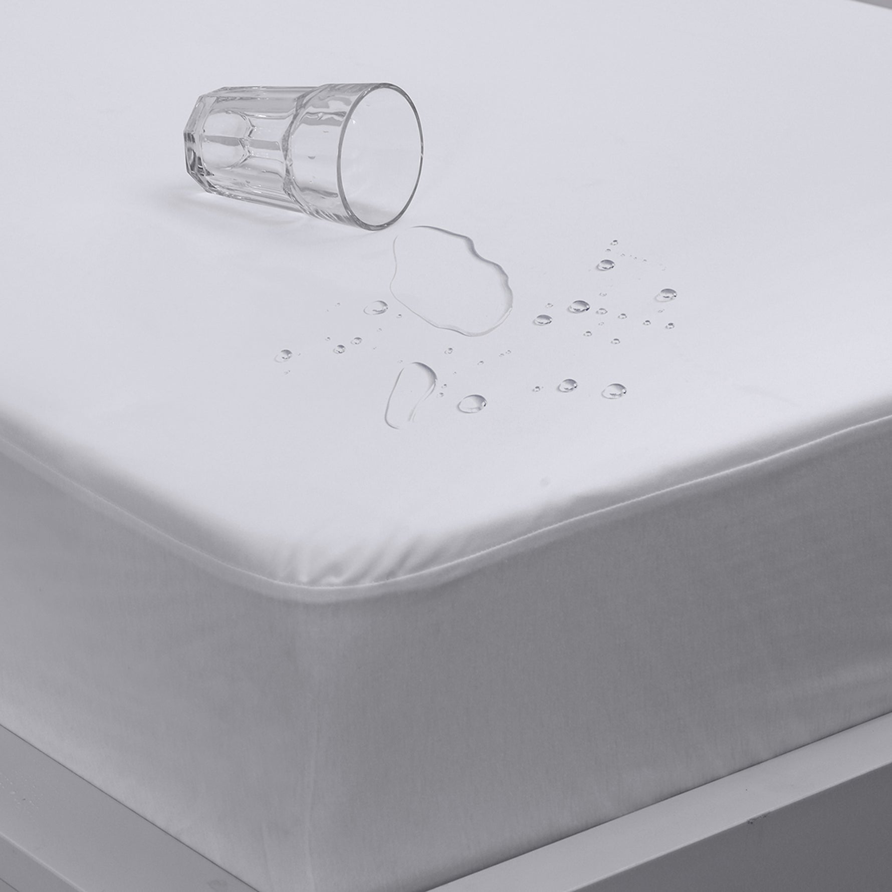 Close-up of a white Eva Clean waterproof mattress protector with water droplets beading on the surface and an overturned glass, fitted over a mattress.
