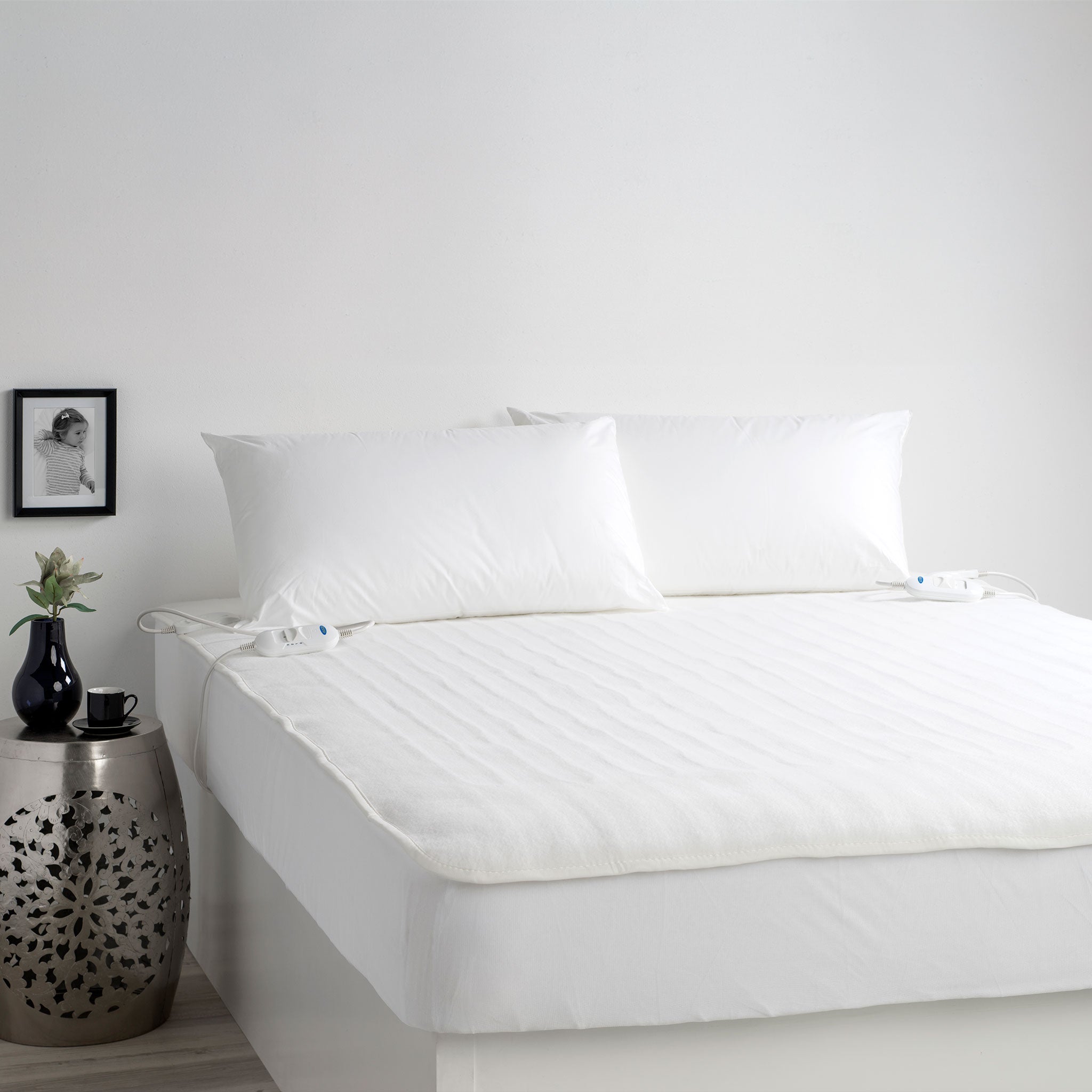 Electric blanket on a bed with two controllers, paired with two plain pillows without covers, offering a warm and comfortable sleep experience. Ideal for cosy nights with adjustable warmth.
