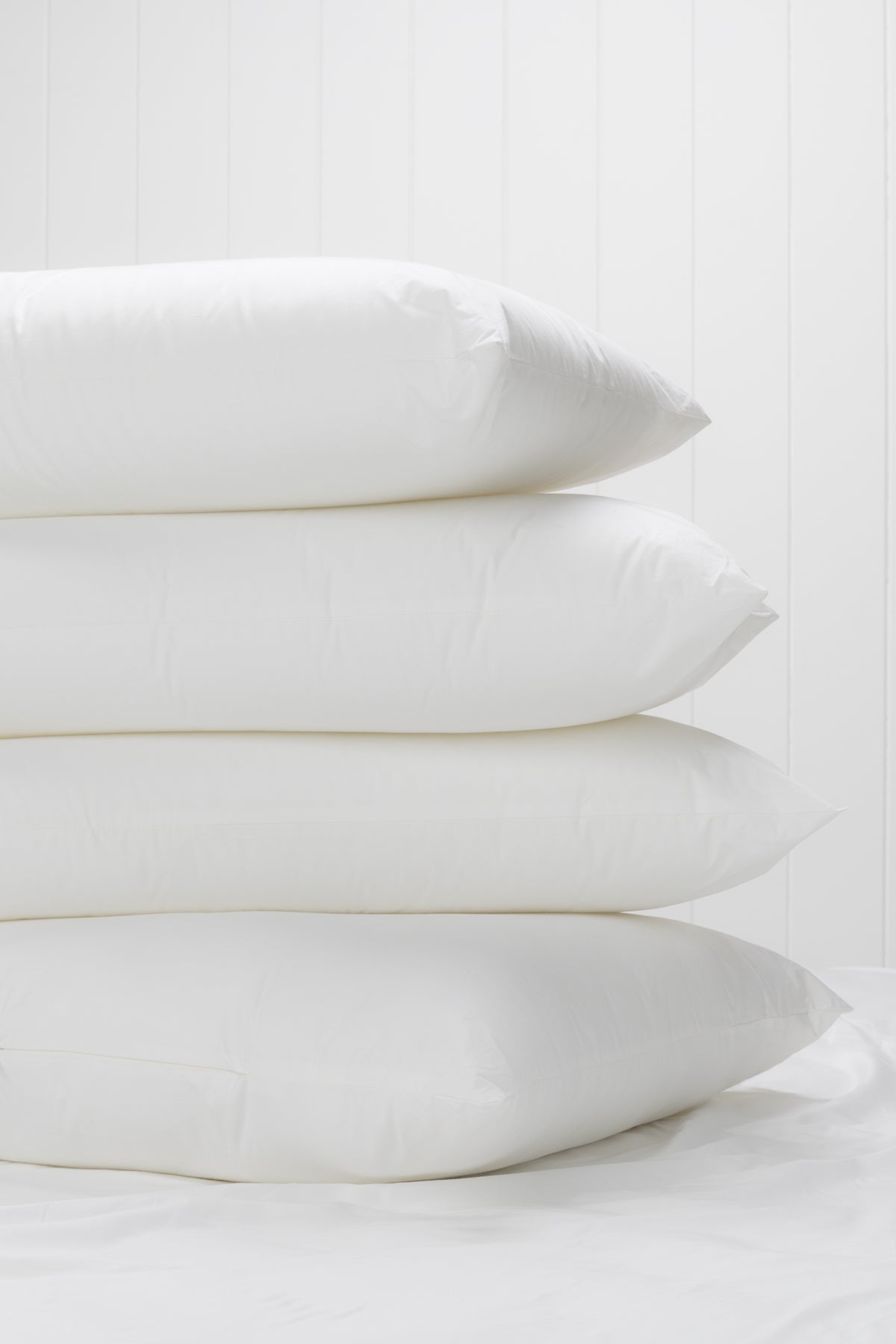 Stack of four white pillows on a bed with a white timber-panel wall in the background.