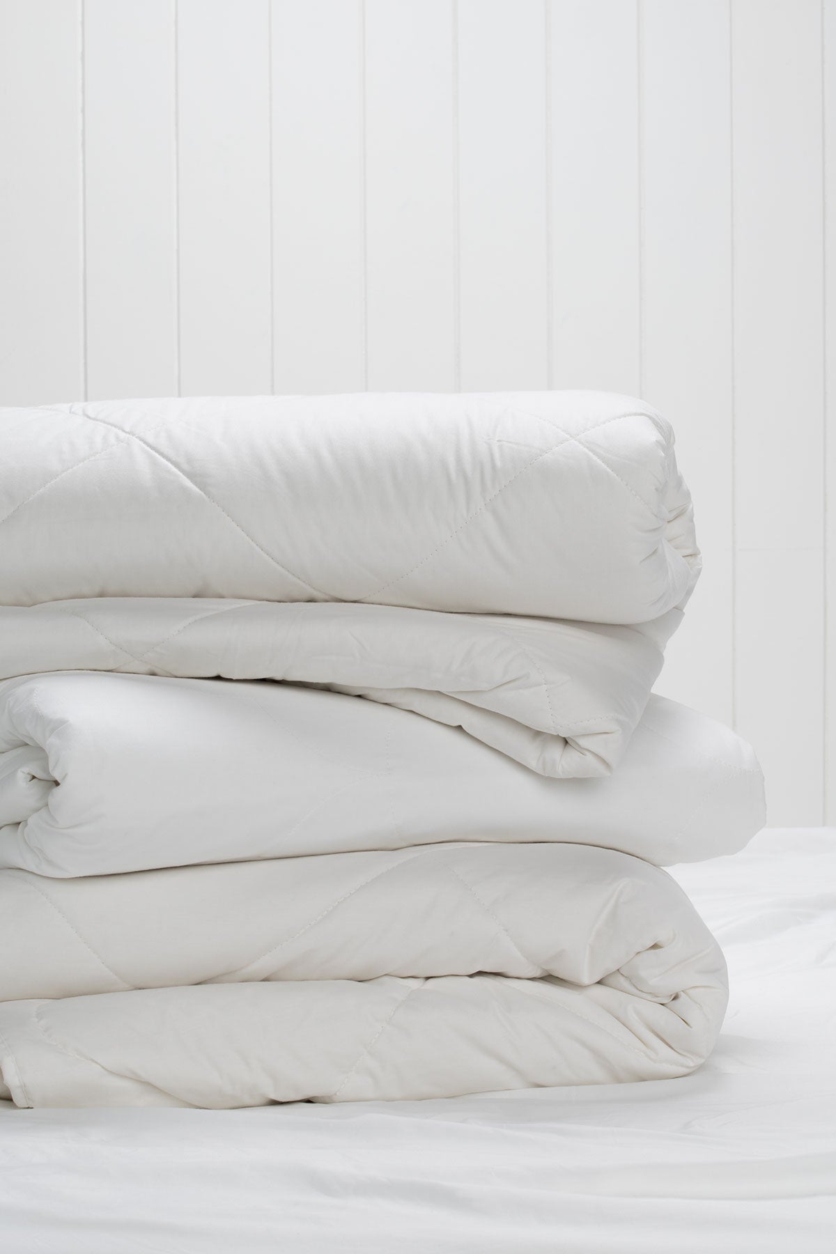 Stack of folded white quilts with diamond quilting on a bed against a white timber-panel wall.