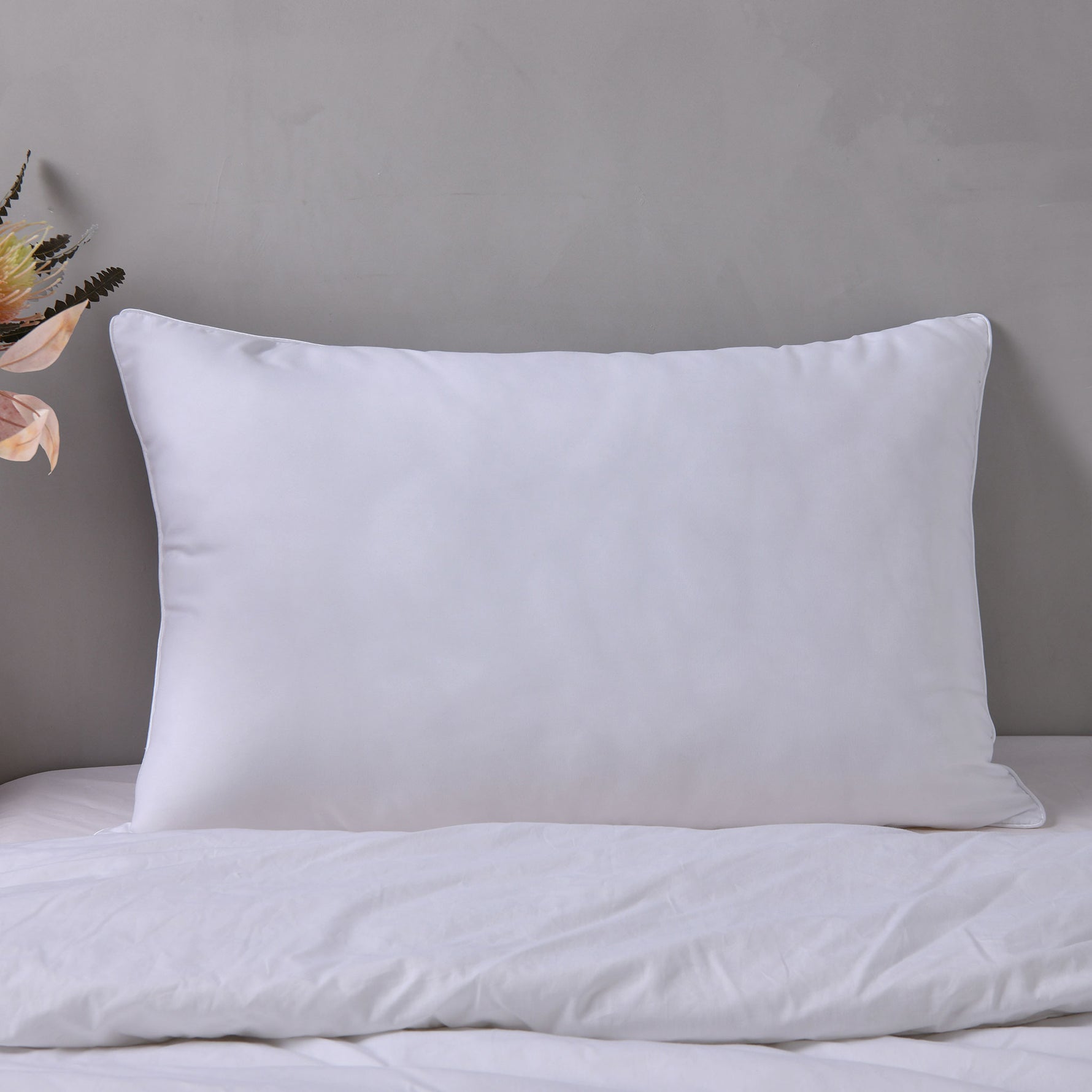 White pillow with piped edging propped upright on a bed against a neutral grey wall.