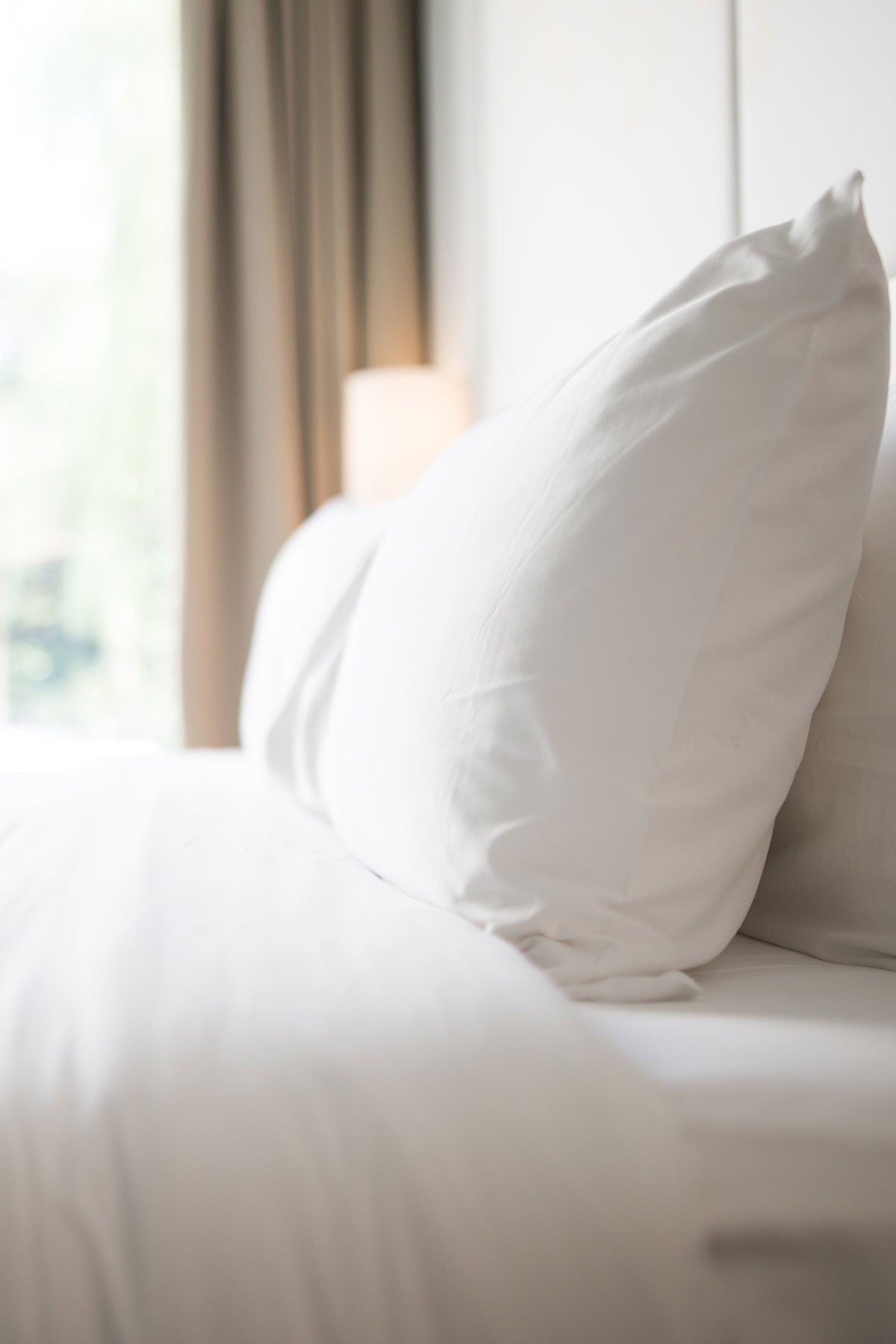 Side view of white Cotton Deluxe cotton percale bed linen with matching pillowcases and a blurred window in the background.